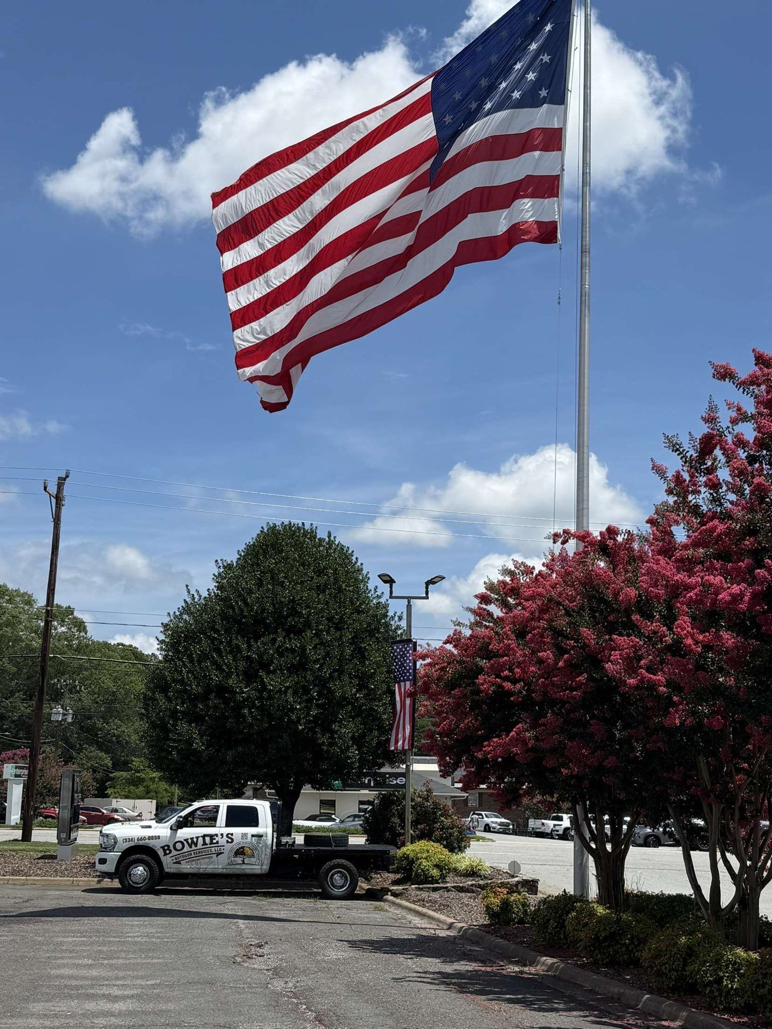 American flag waving high on a pole, with a truck parked in front of trees under a blue sky.