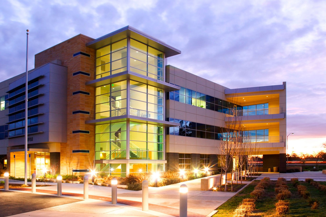 Modern glass-and-brick office building at dusk with lit windows and landscaped walkway.