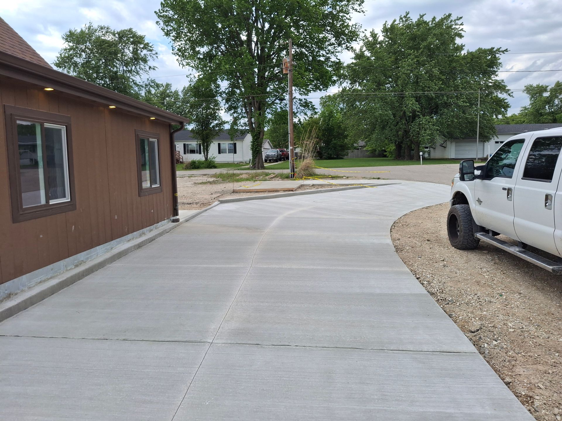 Curved concrete driveway beside a brown building, with a white truck parked on gravel and trees in the background.