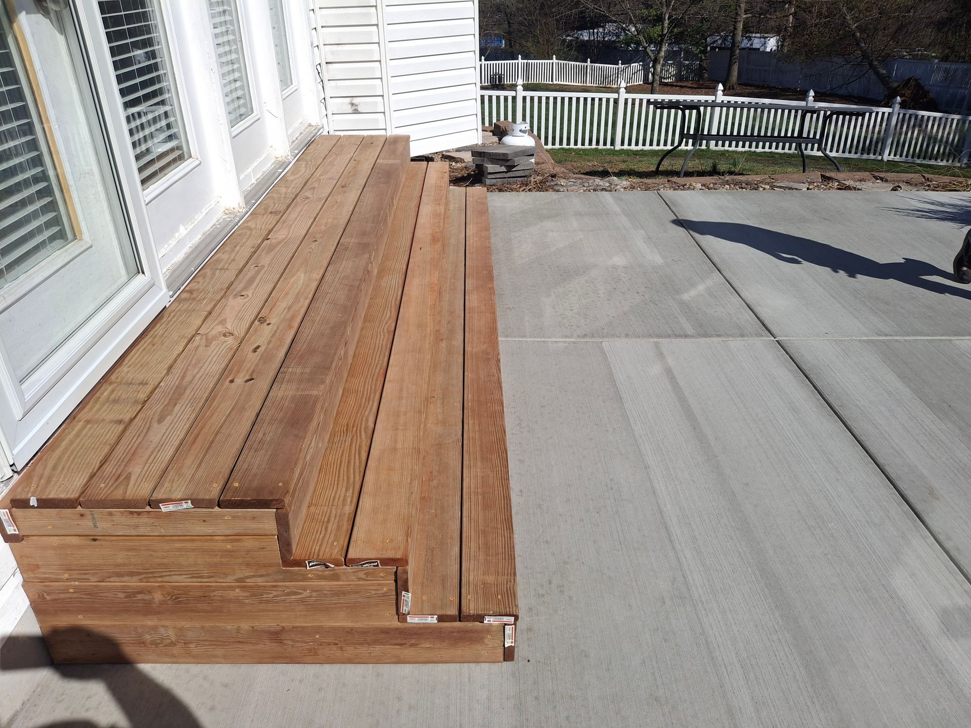 Wooden deck steps beside a house, leading down to a concrete patio and fence.