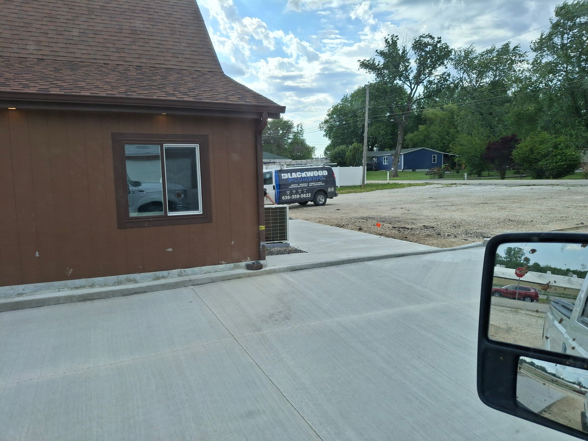 Brown building beside a paved lot, with a parked van, trees, and a side-view mirror in the foreground.