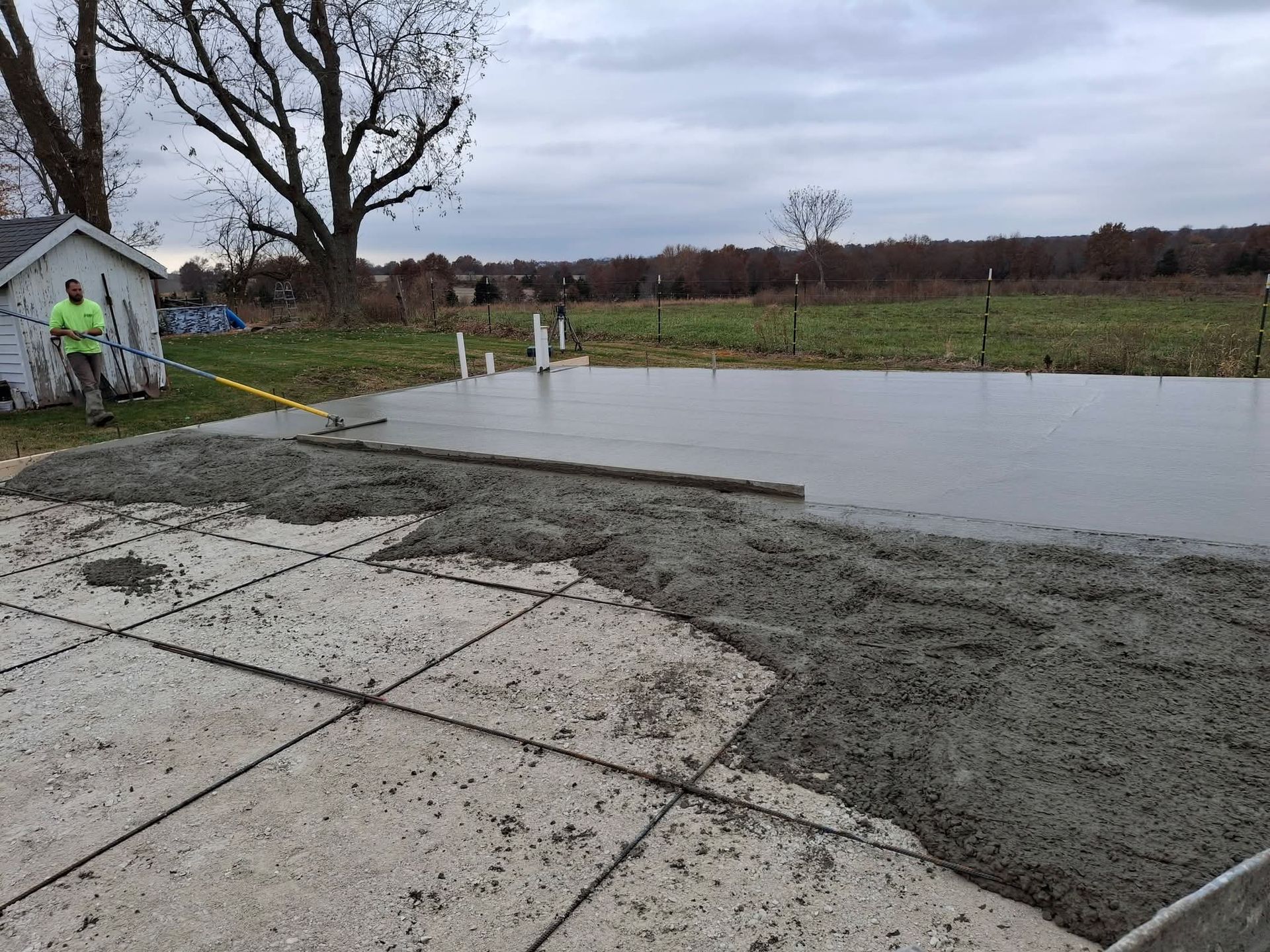 Fresh concrete patio beside a small shed, with workers smoothing a newly poured slab on a cloudy day