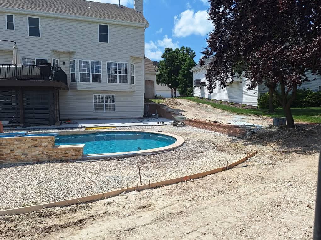 Backyard with a small blue pool beside a house, dirt ground, and a tree under a bright sky