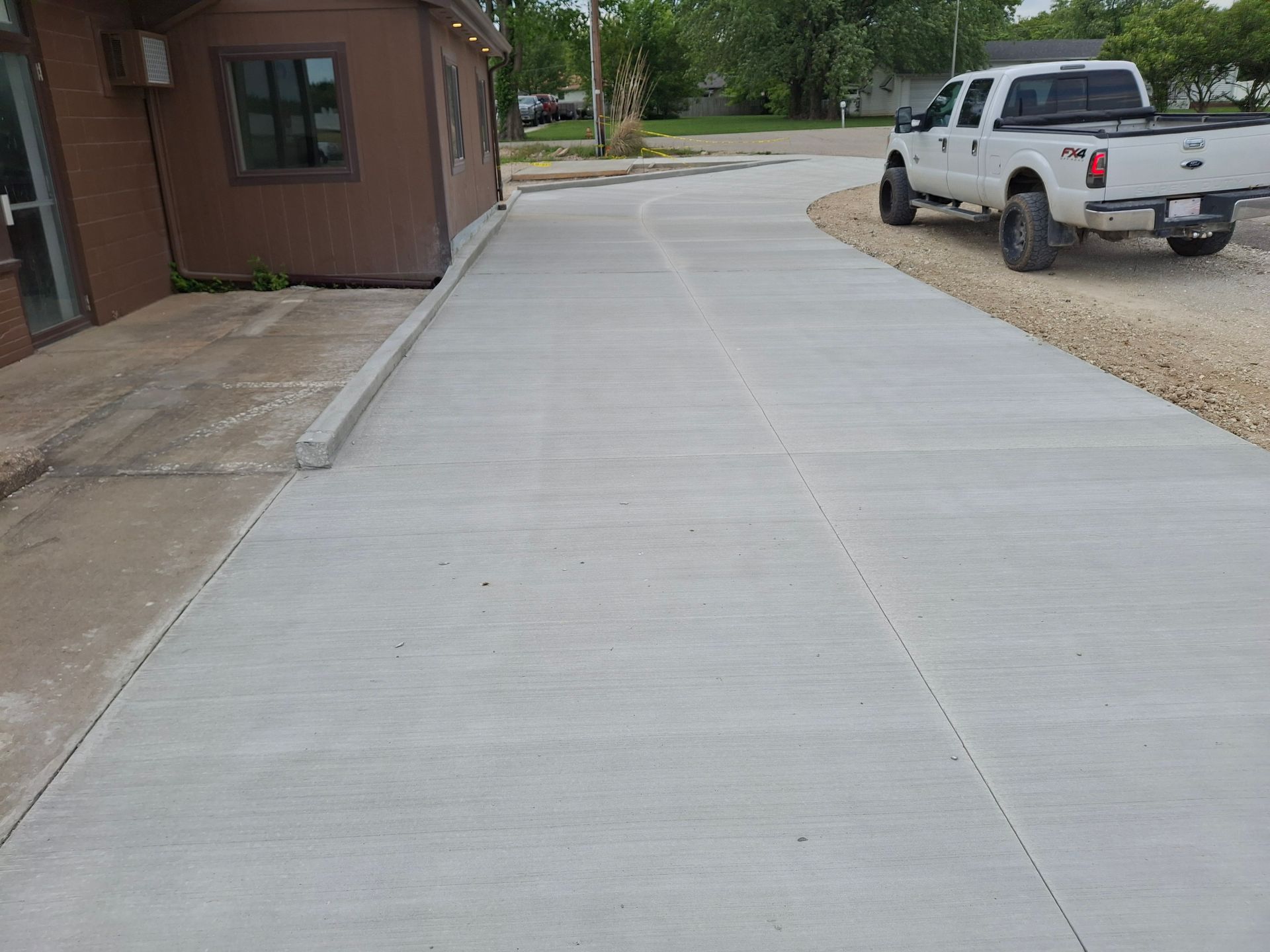 Concrete driveway beside a brown building, with a white pickup truck parked on gravel nearby.