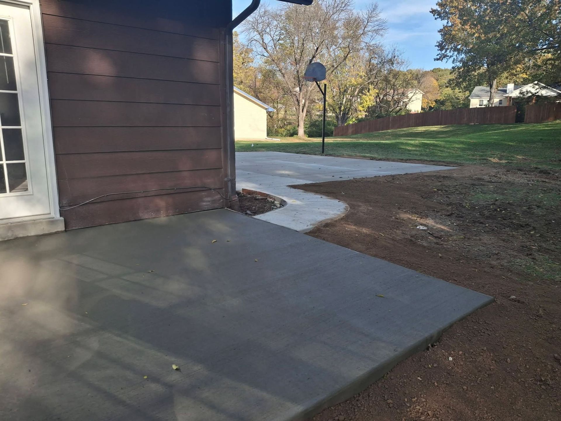 Concrete patio beside a brown building, with a curved sidewalk leading to a basketball hoop in a grassy yard.