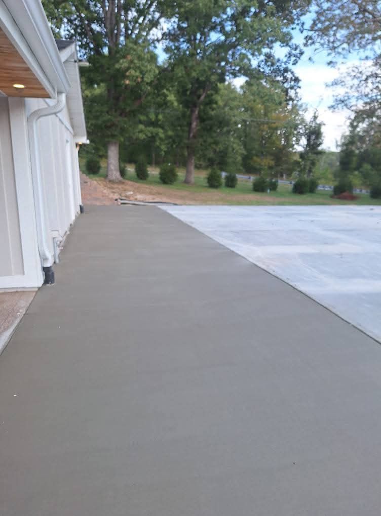 Concrete walkway beside a white building, with trees and a paved driveway in the background.
