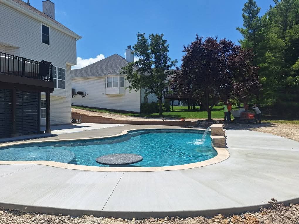 Backyard pool with curved concrete deck beside a house, trees, and a sunny clear sky