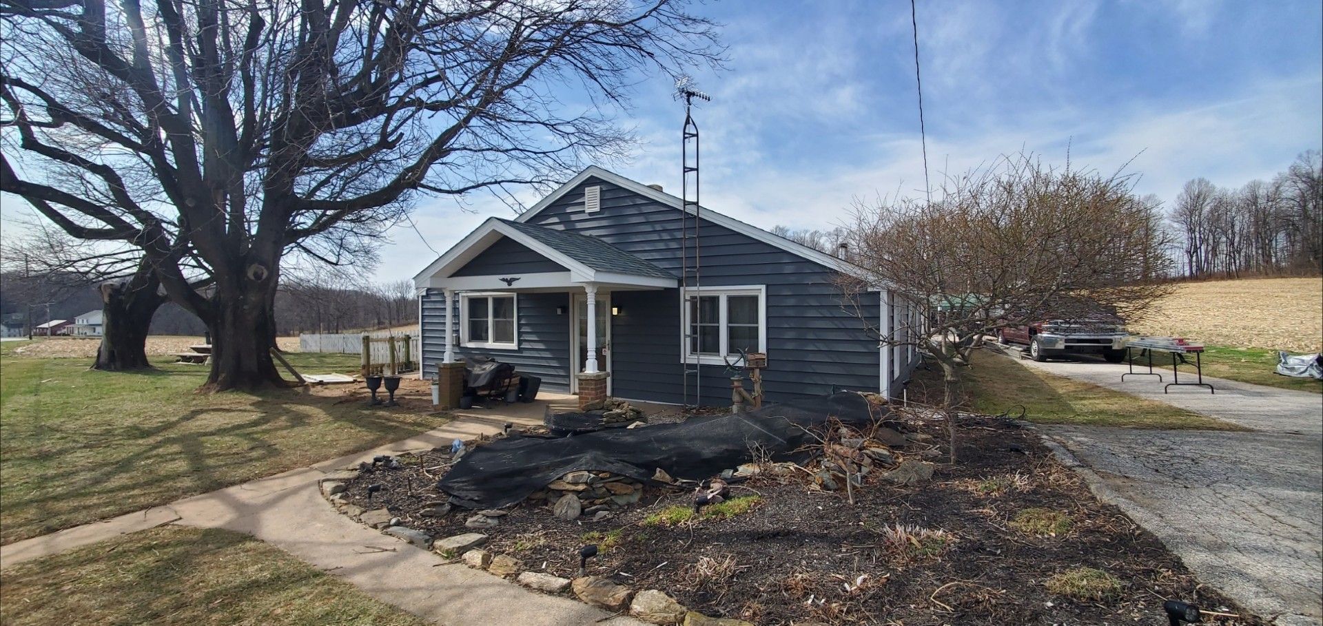 A small, dark gray, single-story house with a white trim porch, set beside a large leafless tree on a sunny day.