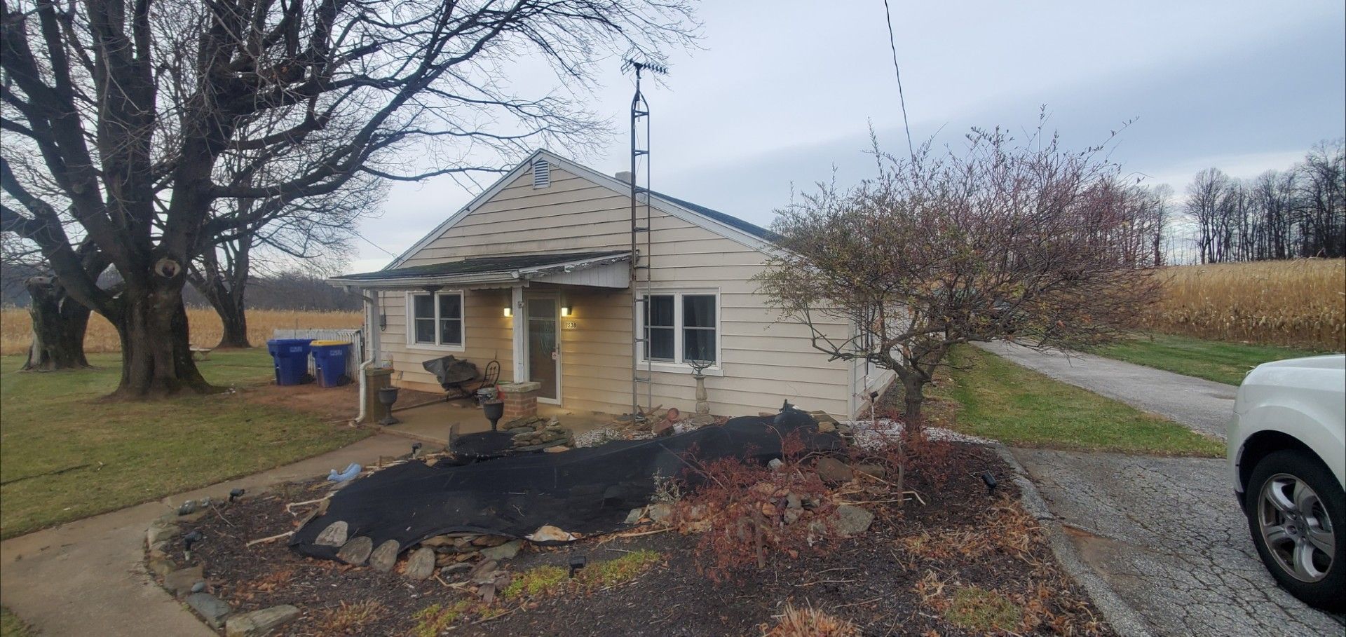 A single-story, light-colored house with a front porch, located in a rural setting with large trees and a field.