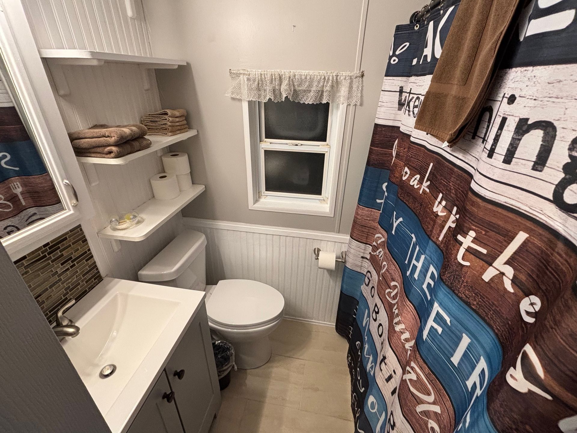 Small bathroom with a toilet, sink, and shower curtain featuring rustic design. White shelves hold towels and toiletries.