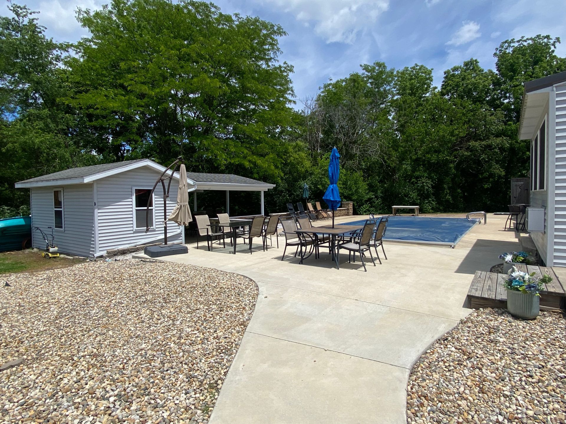 Backyard patio with pool, outdoor dining set, blue umbrella, and small shed under a sunny sky
