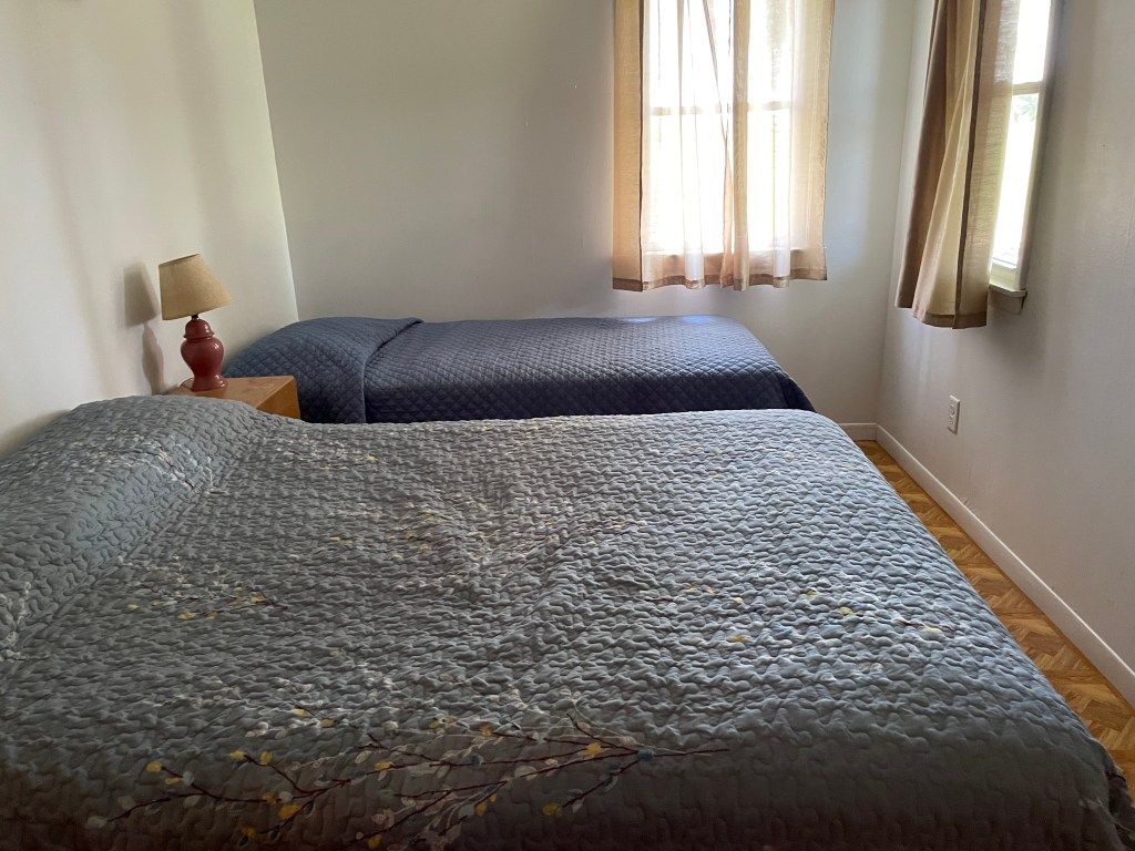 Bedroom with two beds, gray bedspreads, a bedside lamp, and windows with curtains.