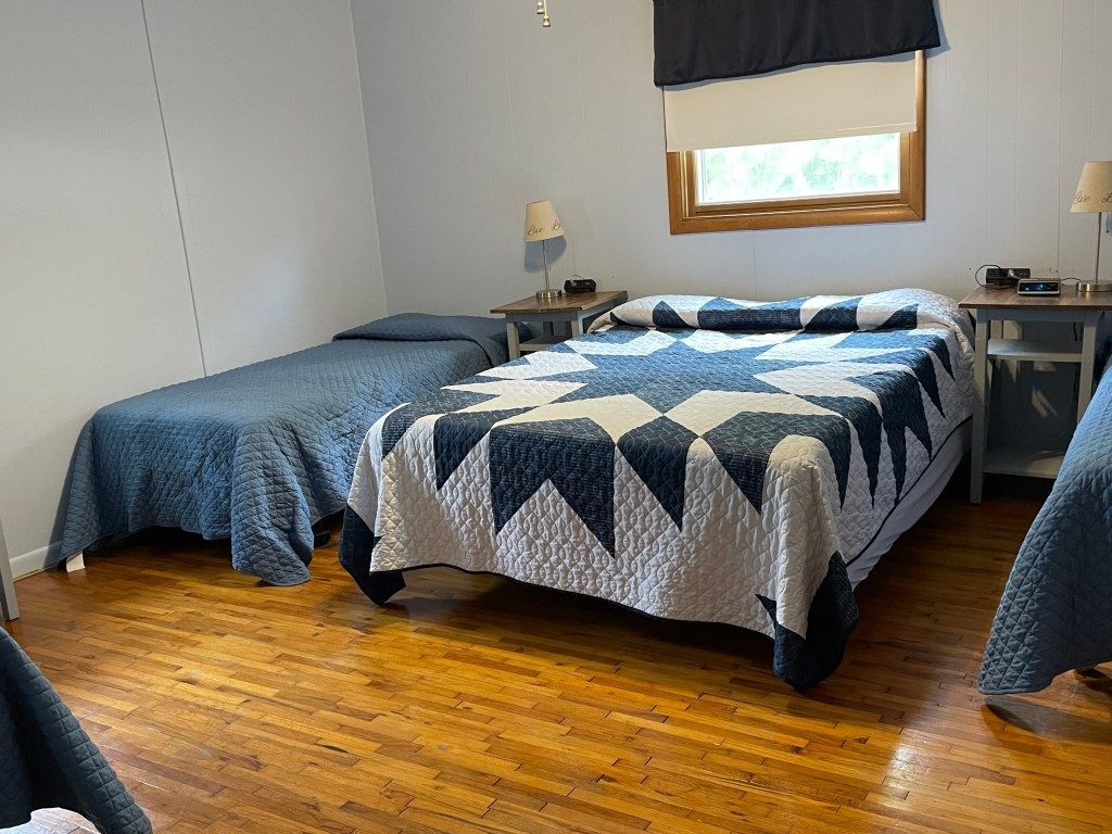 Bedroom with two beds, blue quilts, wood floor, and a window with dark curtain.