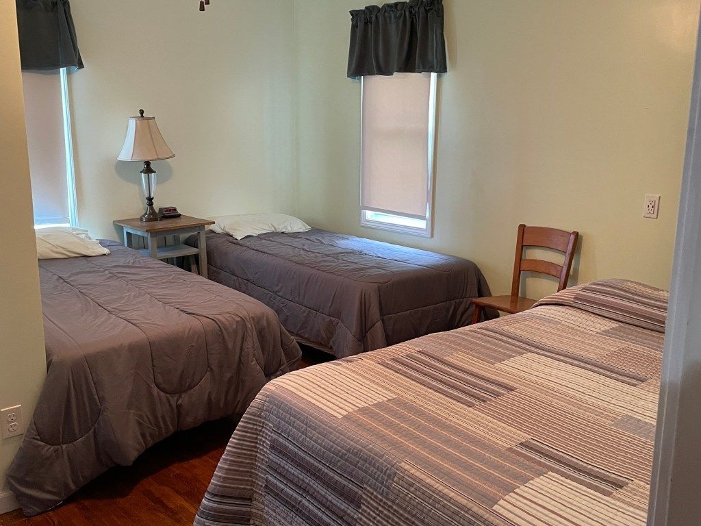Bedroom with three made beds, two windows, a lamp, and a wooden chair on the right.