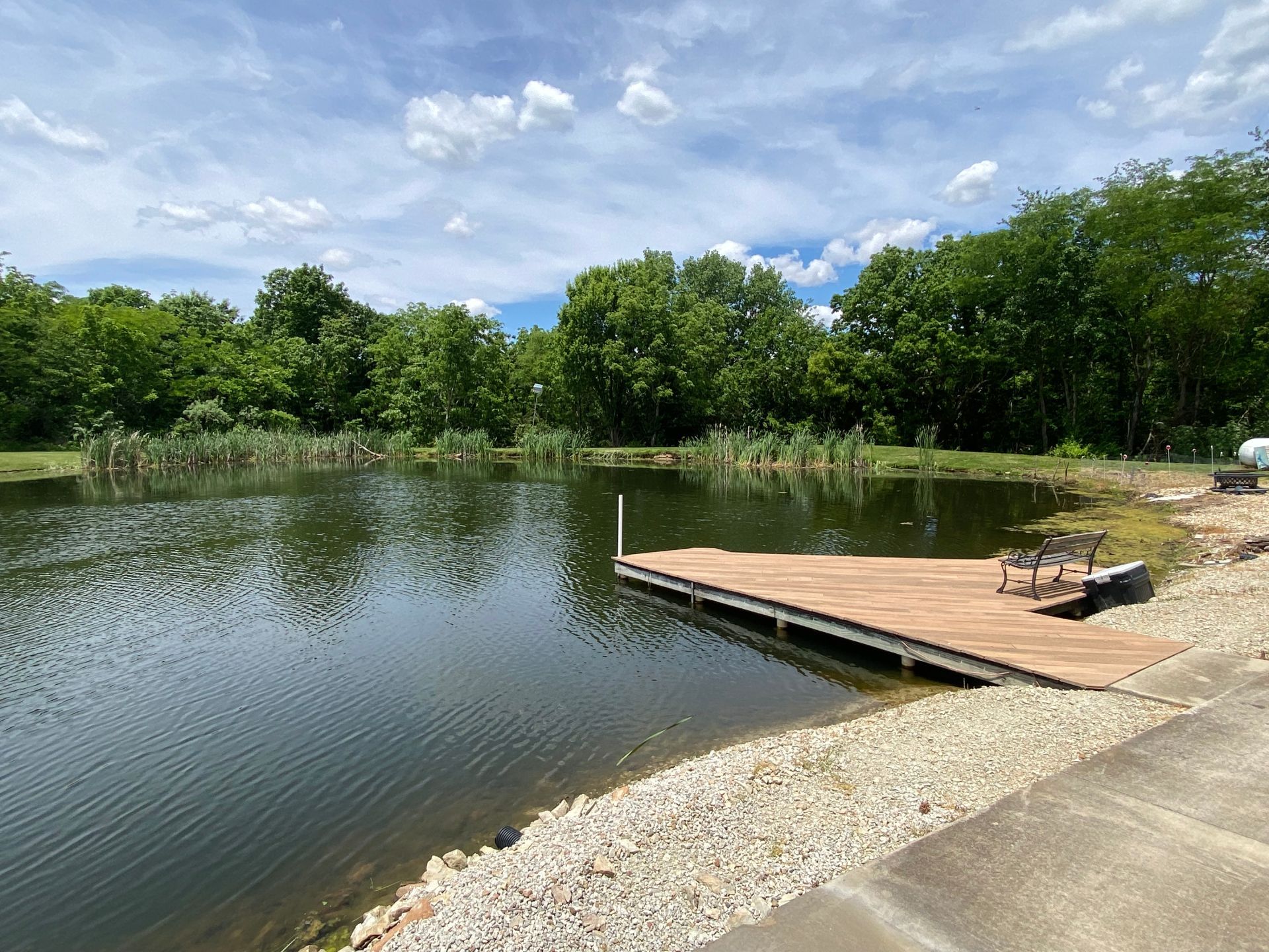 Calm lakeside with a wooden dock, rocky shore, and green trees under a partly cloudy sky