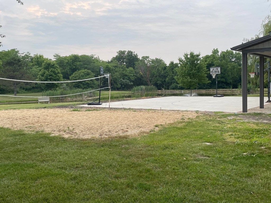 Outdoor sand volleyball court and basketball hoop beside a grassy park area under a cloudy sky