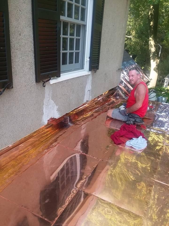 A man is sitting on a copper roof next to a window.