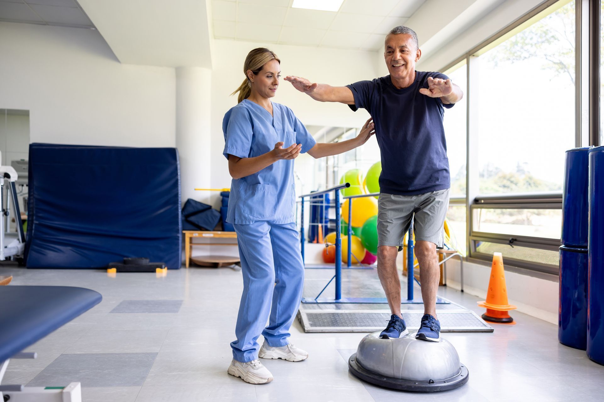 Woman in blue scrubs assists a smiling older man balancing on a dome-shaped exercise ball in a rehab clinic.