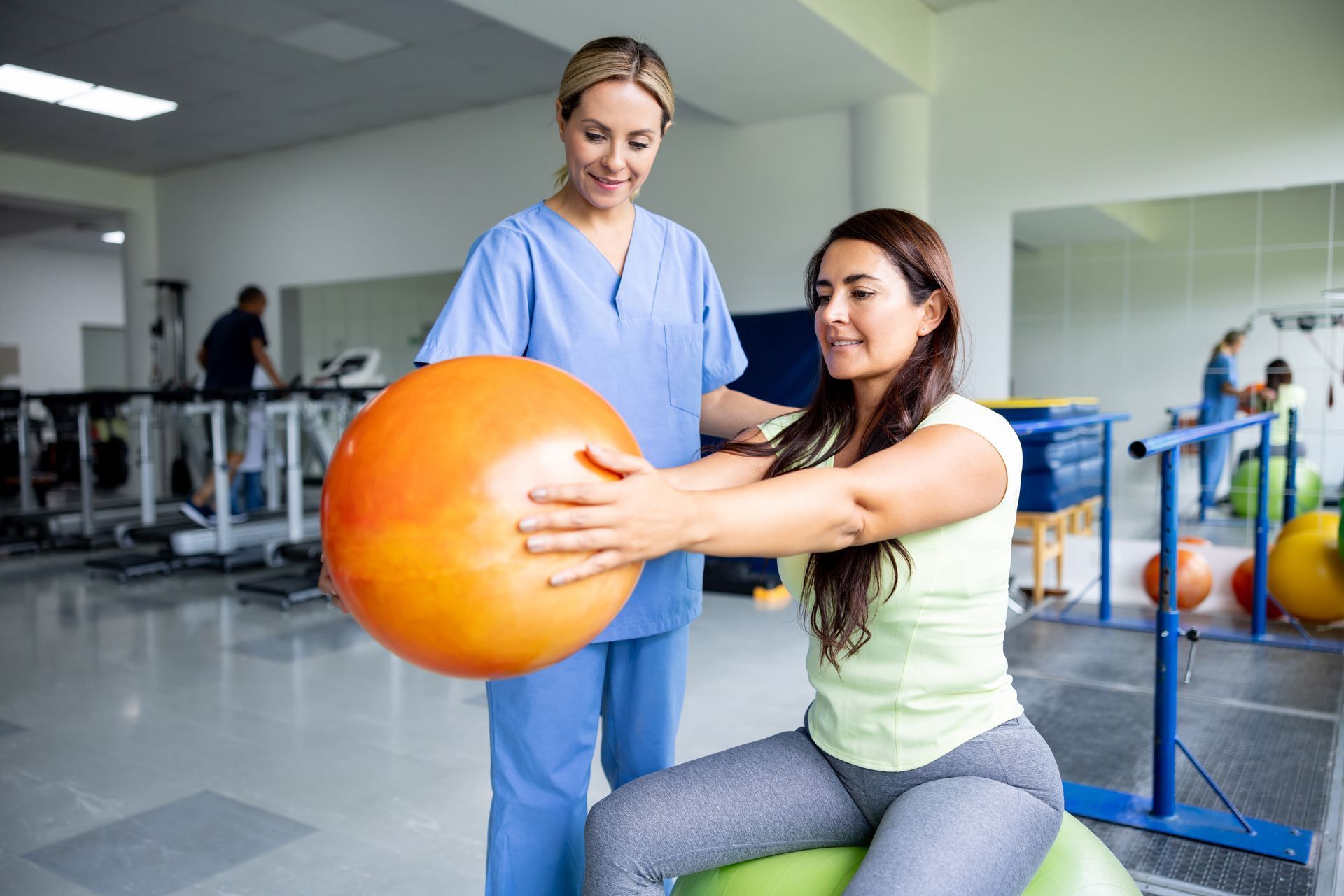 Woman exercises with an orange ball, assisted by a physical therapist in a light-filled gym.