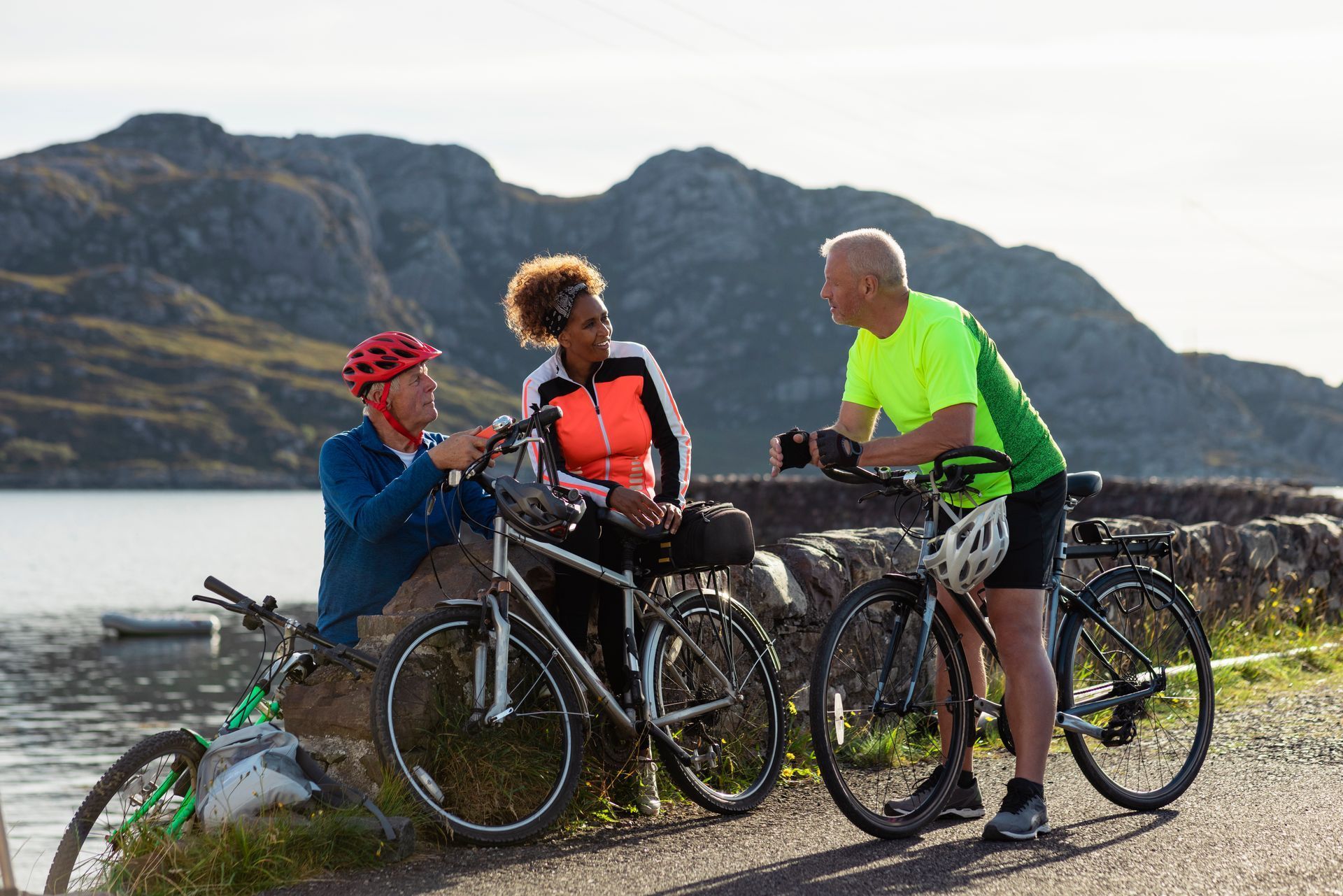 Three cyclists, overlooking a lake and mountains, talking and resting with their bikes.