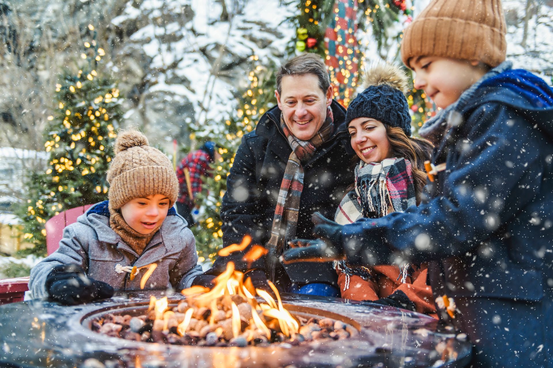Family roasting marshmallows over a fire pit outdoors in winter; snow falling, festive lights.