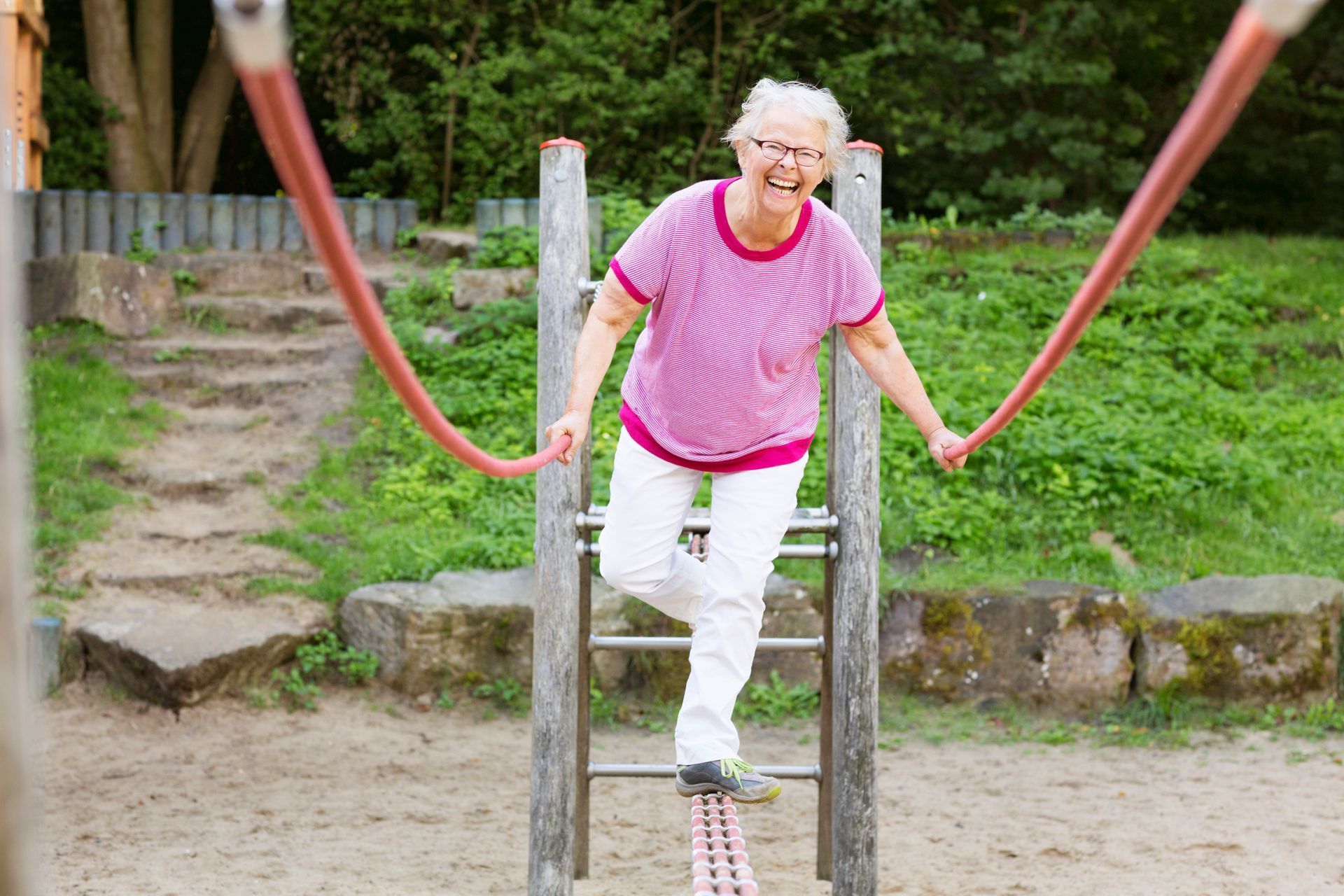 Smiling senior woman on a playground balance beam, holding support ropes, white pants, pink top.