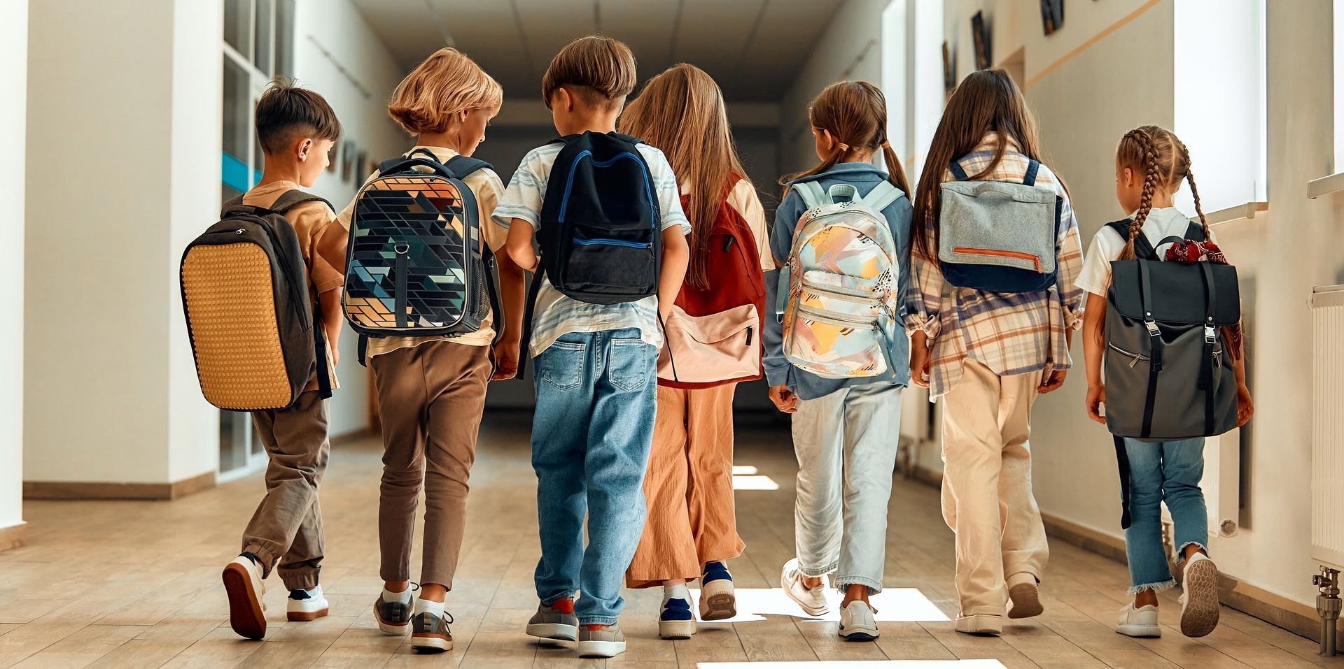 Group of school children walking down a hallway, all wearing backpacks.