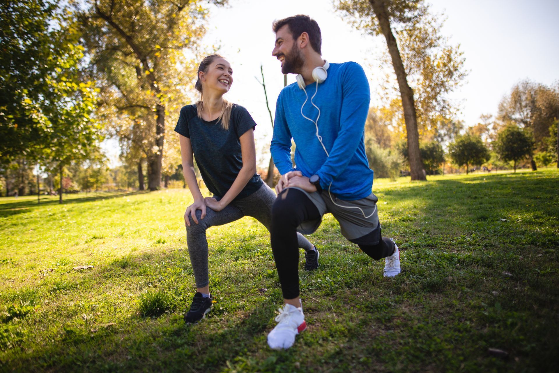 Woman and man smiling, stretching legs in a sunny park. He wears headphones, she laughs, both in workout clothes.