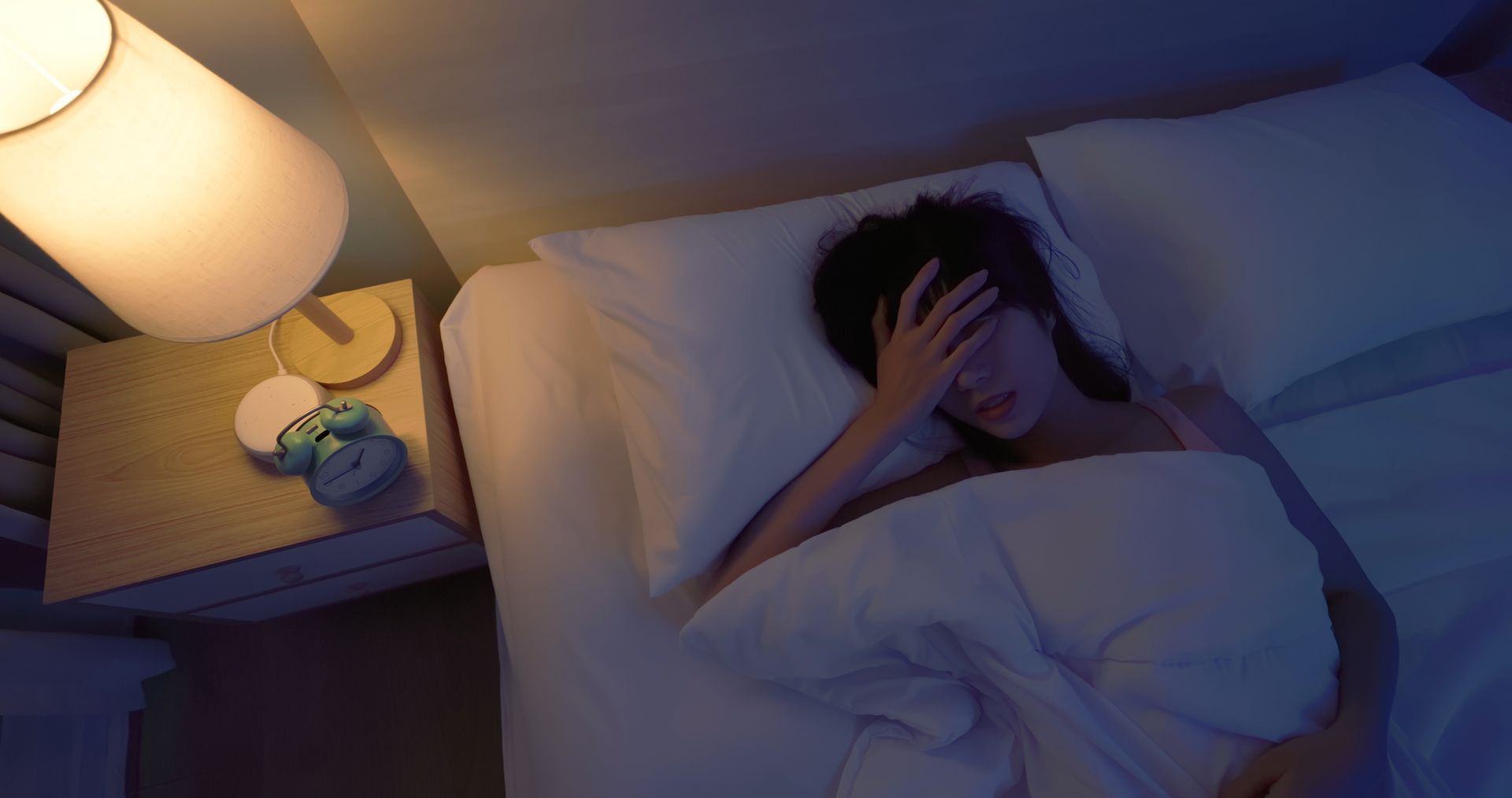 Woman in bed, hand on forehead, illuminated by bedside lamp, appears to have trouble sleeping.