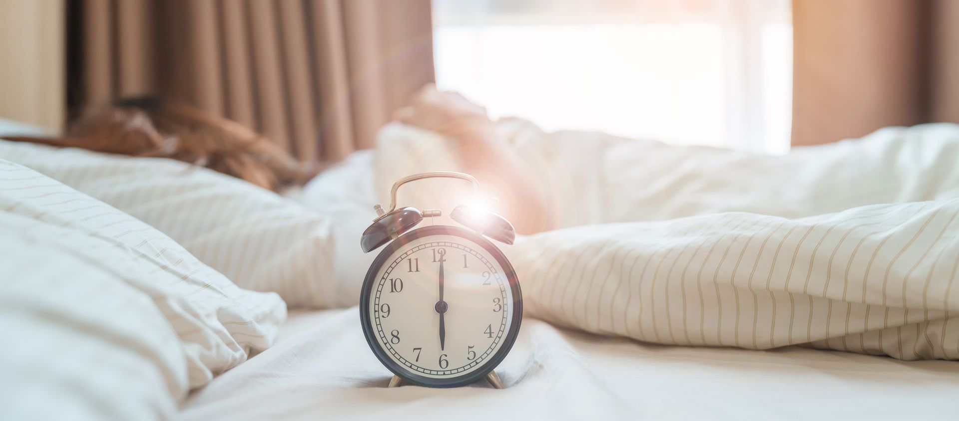 Alarm clock on bed with a person asleep under the covers, daylight streaming in from the window.