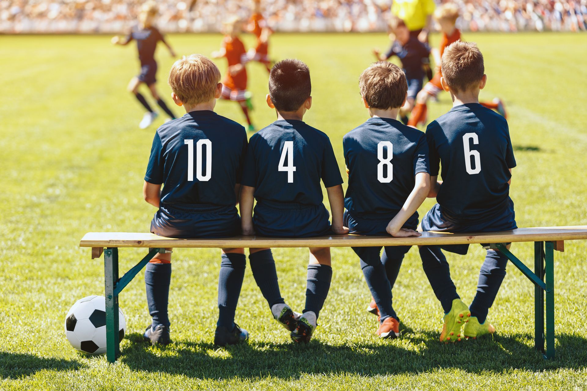Four young soccer players, in blue jerseys with numbers, sit on a bench watching a game on a sunny field.
