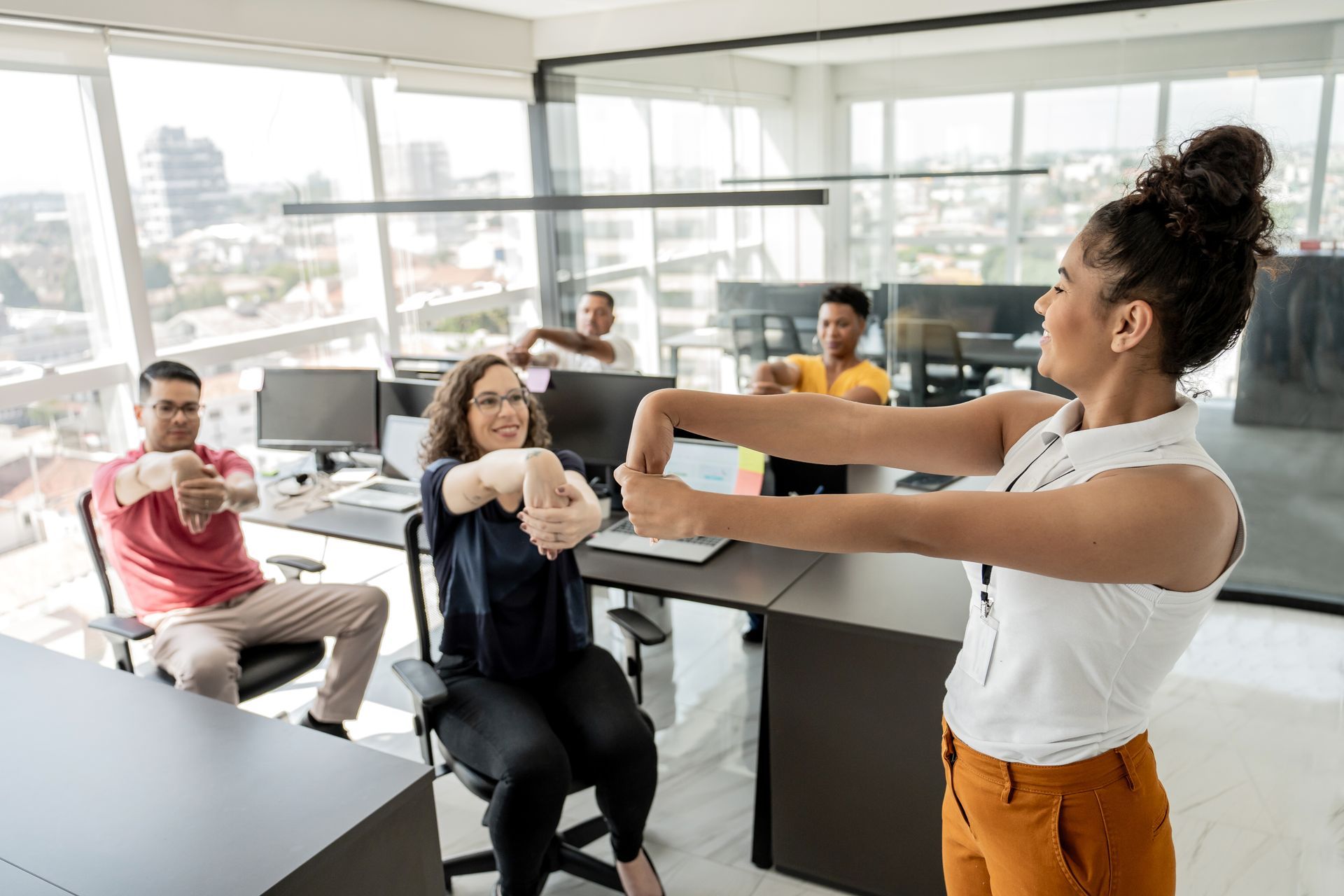 Office workers stretching during a work break. Group led by a woman in a white top, with city views.