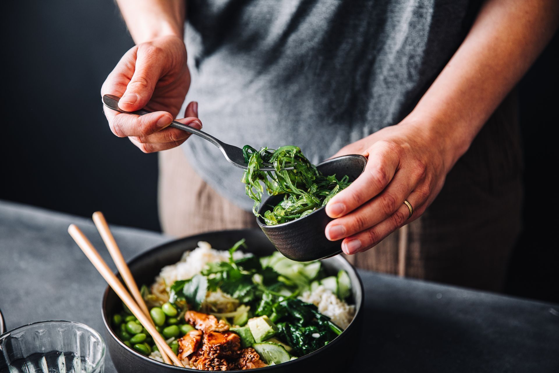 Person adding seaweed salad to a poke bowl with chopsticks and side of edamame.