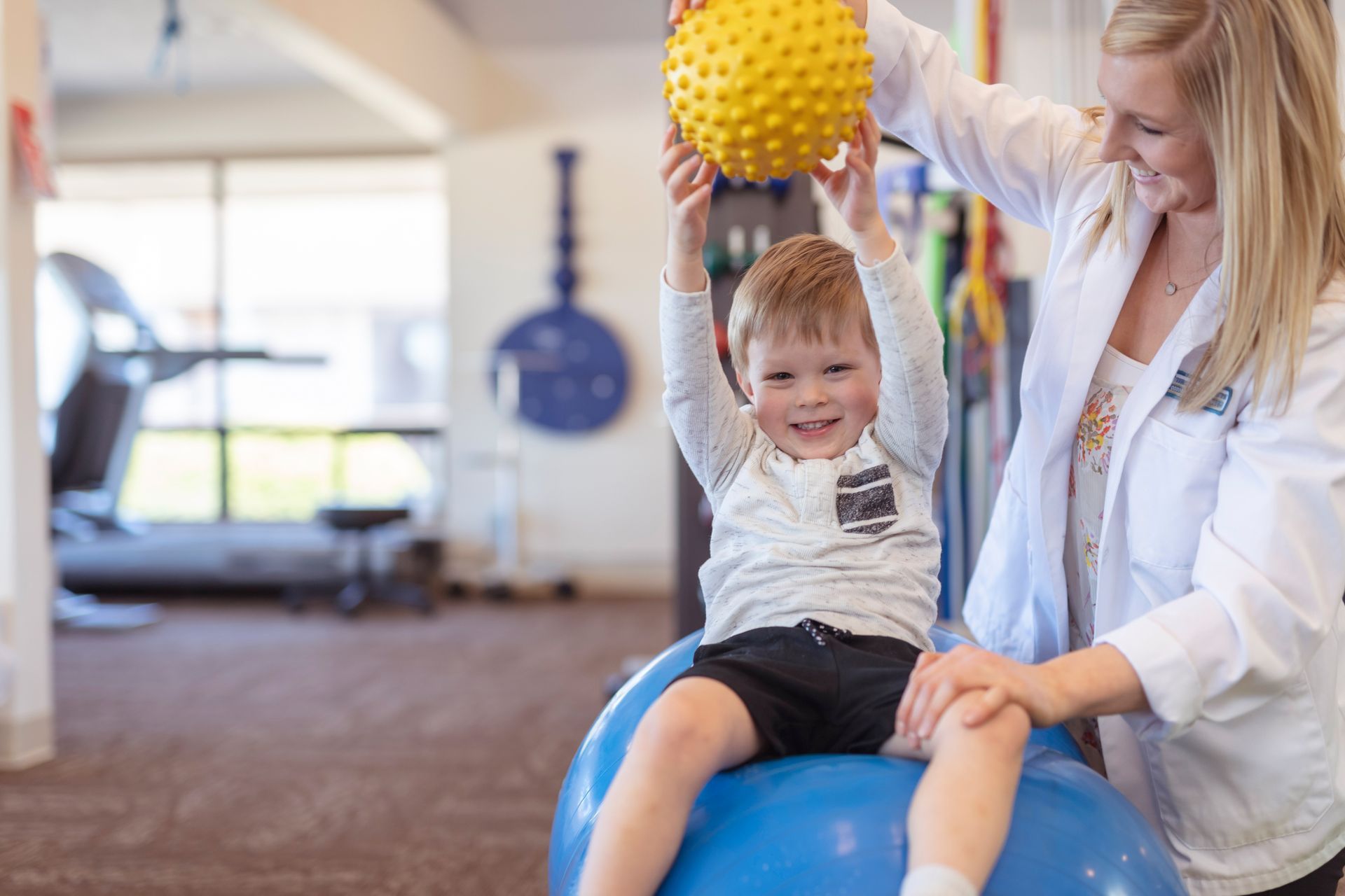 Woman helps a smiling child on a blue exercise ball raise a yellow spiky ball in a therapy room.