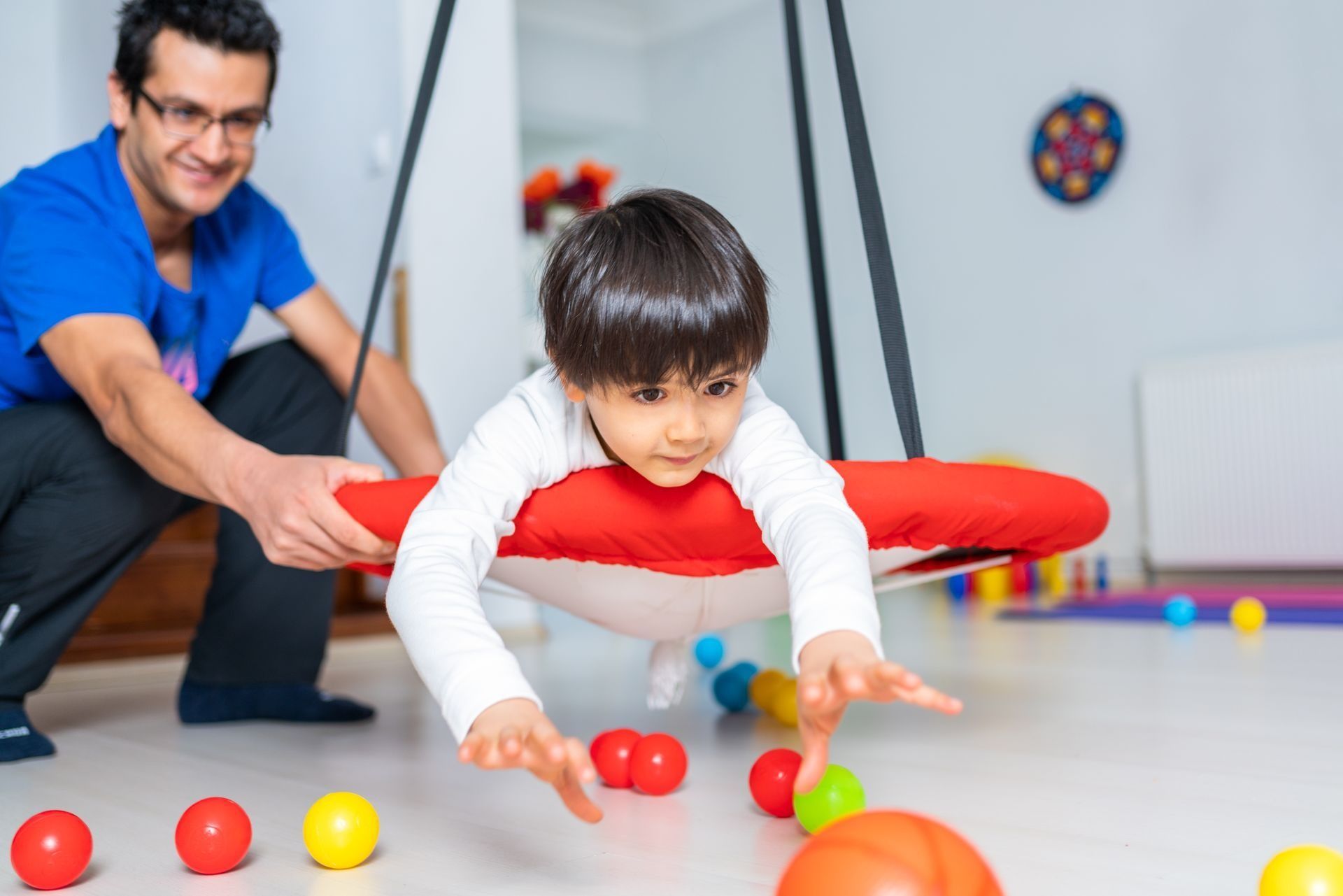 A child on a red swing reaches for colorful balls on the floor, supervised by a smiling adult.