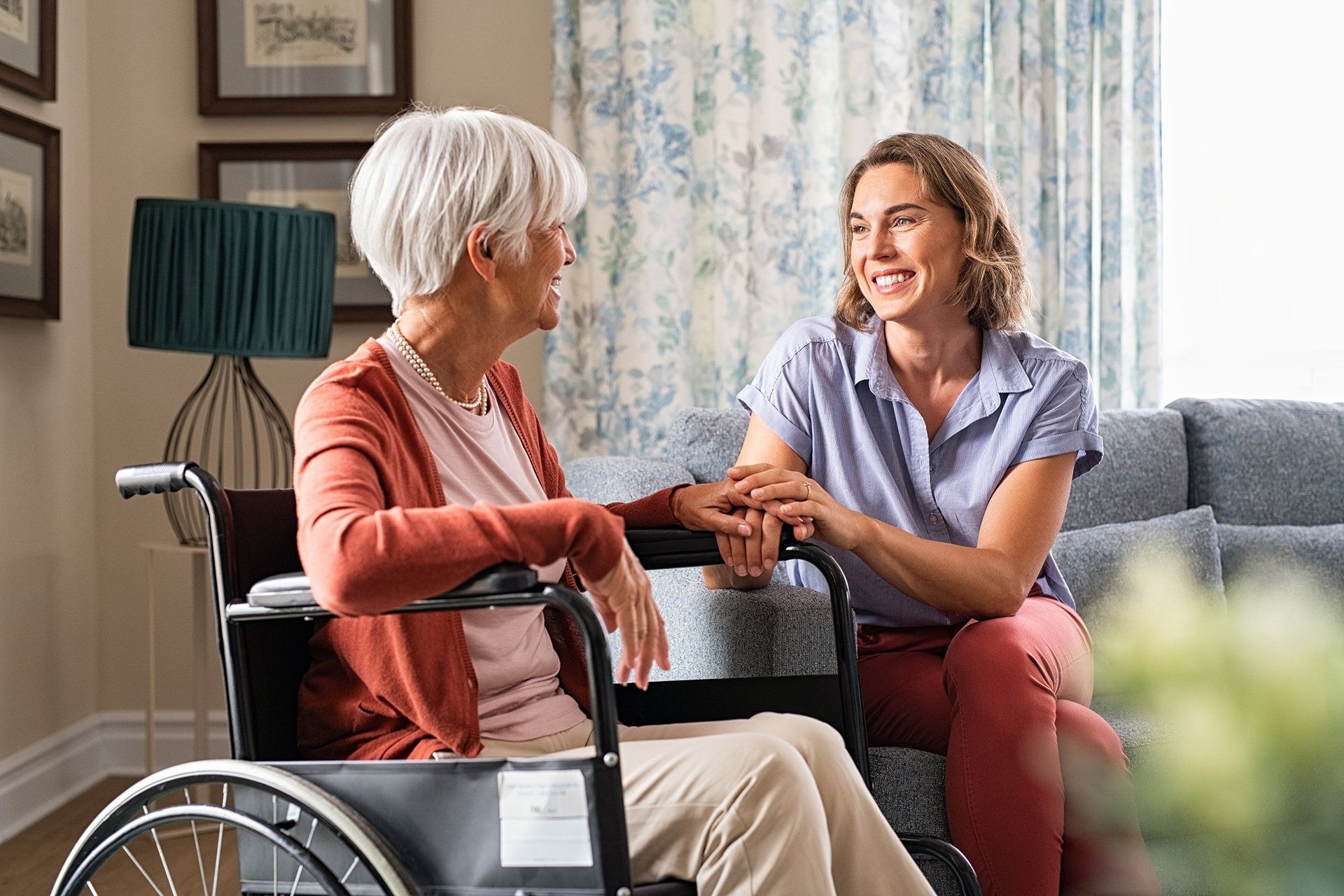Woman in wheelchair and younger woman smiling, holding hands, in a living room.