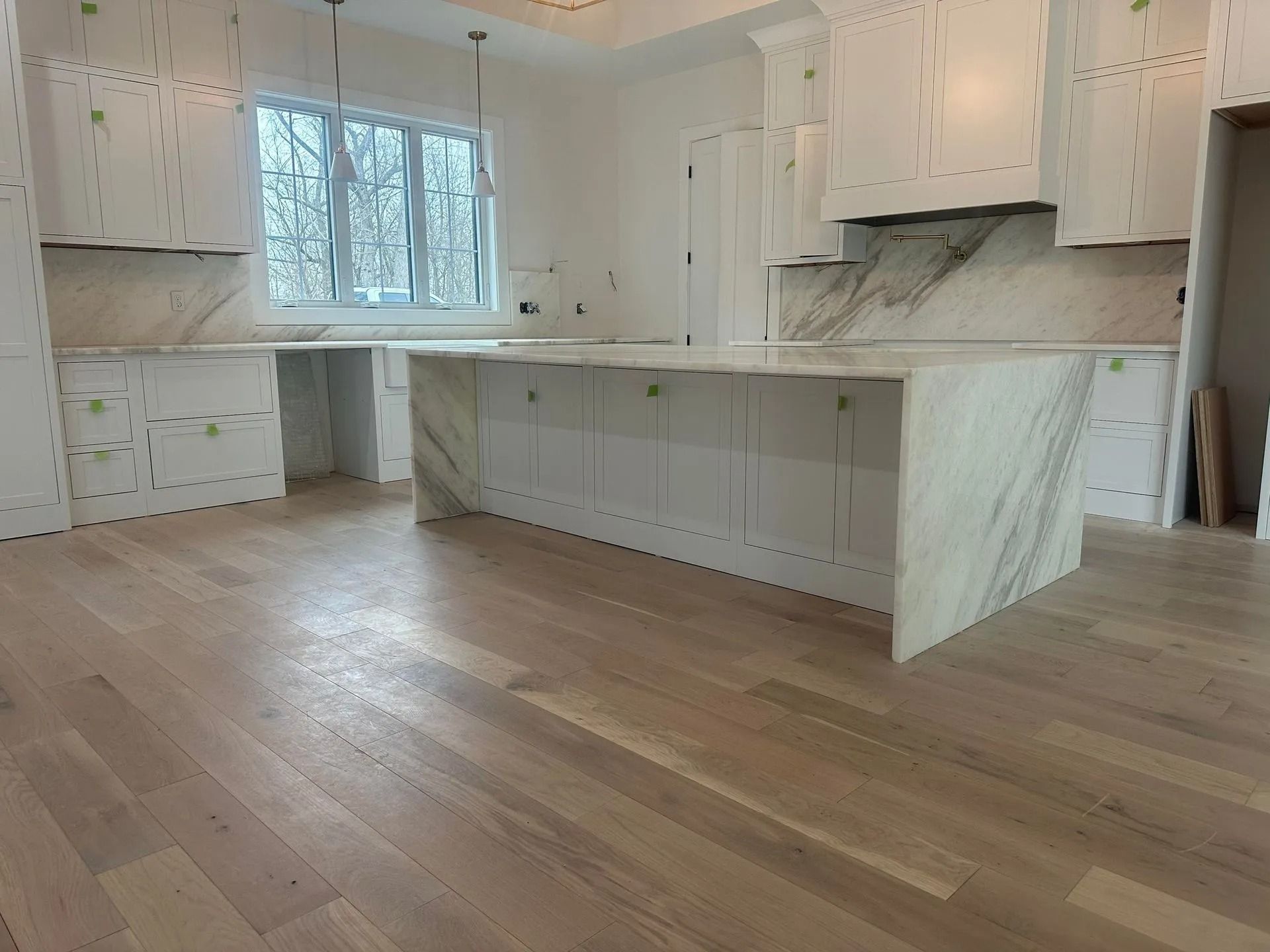 Newly built kitchen with white cabinets and marble countertops. Wooden floors and large window.