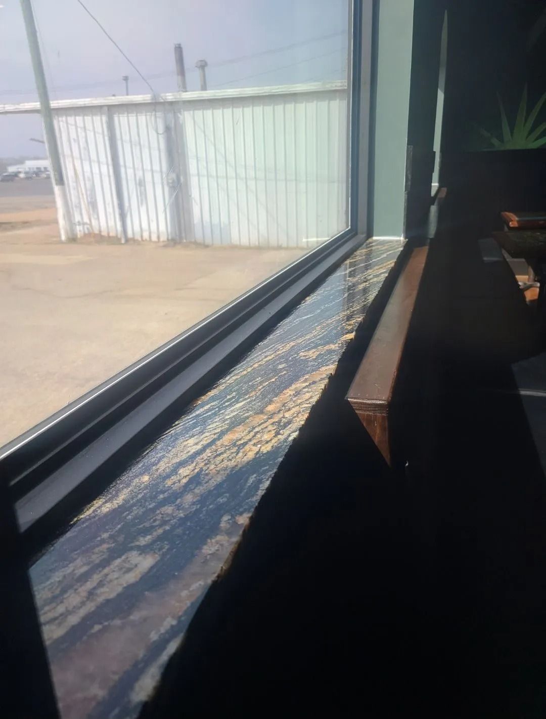A window sill with a reflective, mottled surface, next to a window overlooking a sunny outdoor area.