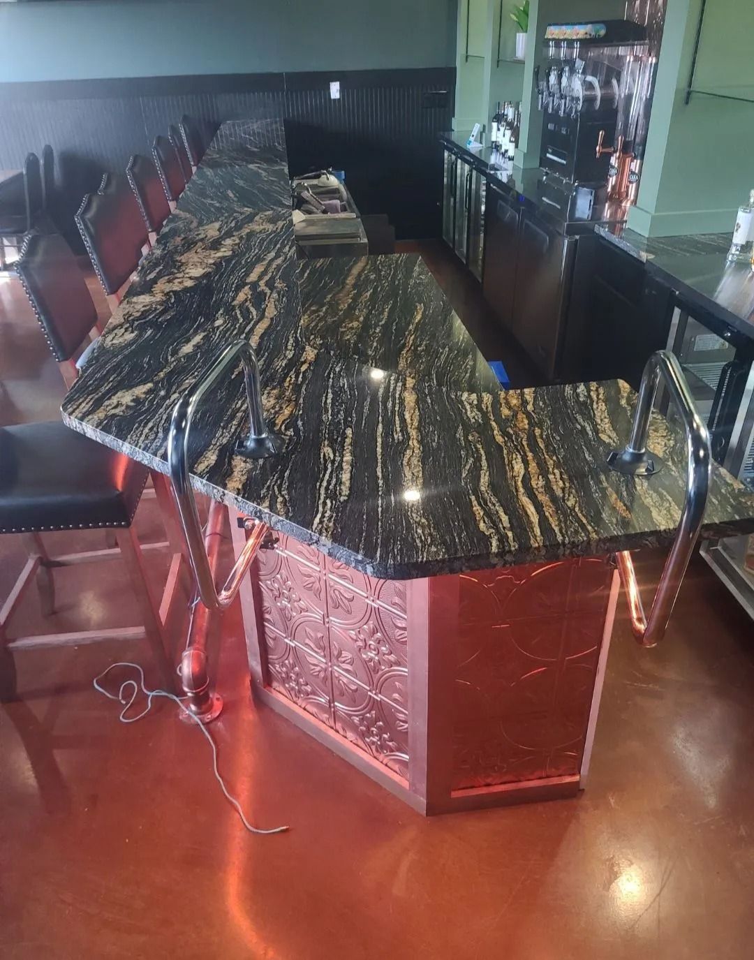 Bar with black granite top and copper-toned facade. High bar stools. Reflected in shiny floor.