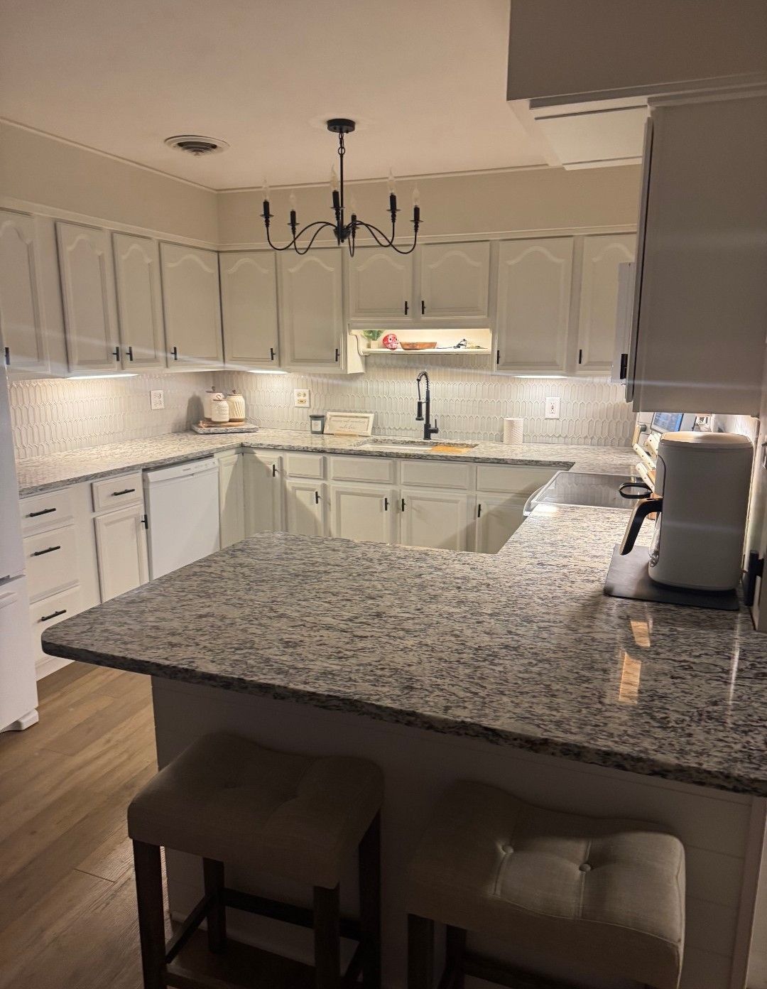 White kitchen with granite countertops, black fixtures, and a chandelier. Two beige bar stools.