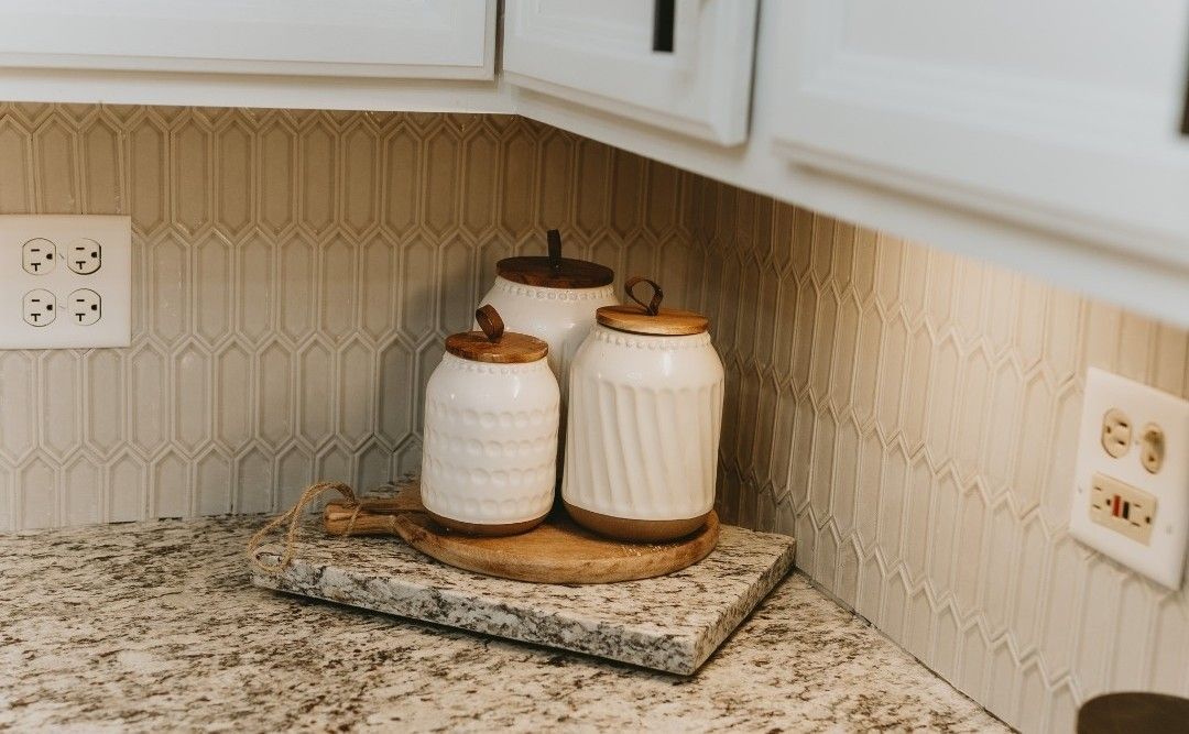White ceramic canisters with wooden lids on a wooden board atop a granite countertop in a kitchen.