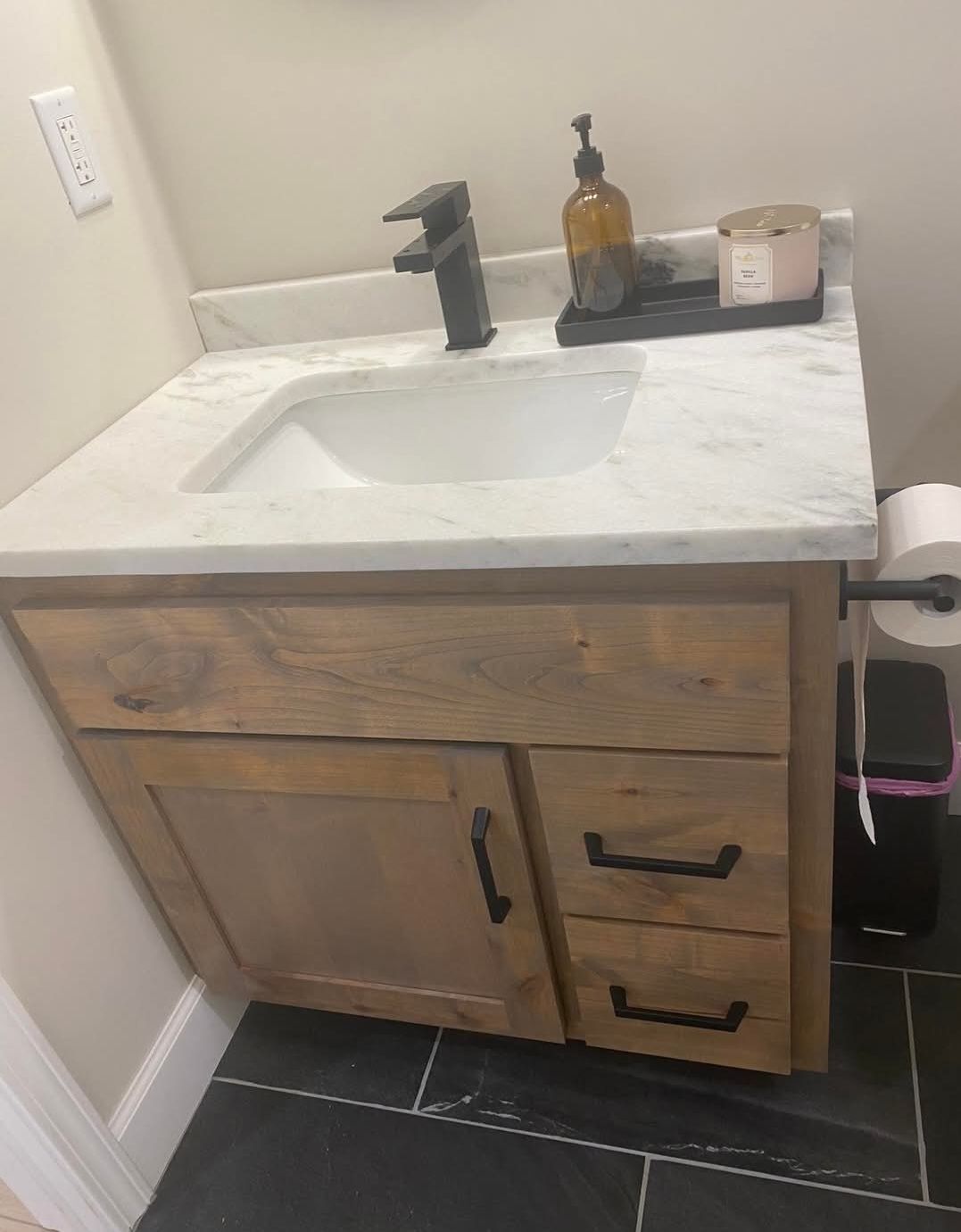 Bathroom vanity with a wooden cabinet, marble countertop, black faucet, and accessories.