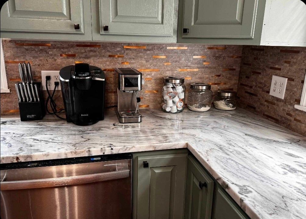 Kitchen countertop with appliances: Keurig, coffee maker, knife block. Backsplash is a mosaic. Green cabinets.