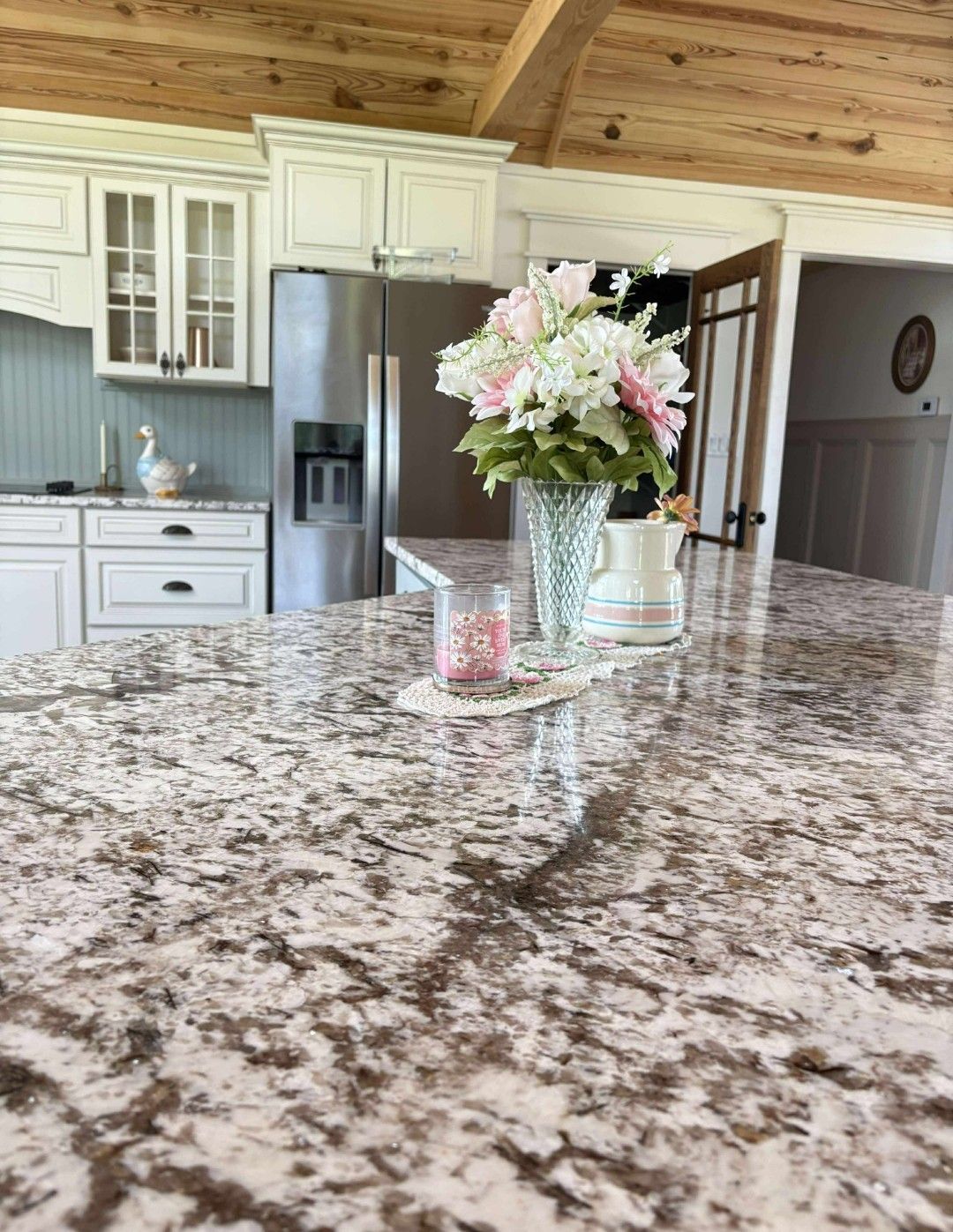 Kitchen island with granite countertop, floral arrangement, stainless steel refrigerator, white cabinets, and wooden ceiling.