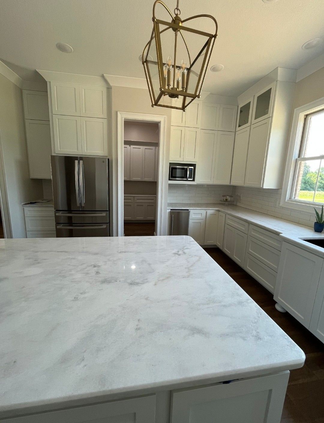 White kitchen with marble island, stainless steel fridge, gold chandelier, and view into pantry.