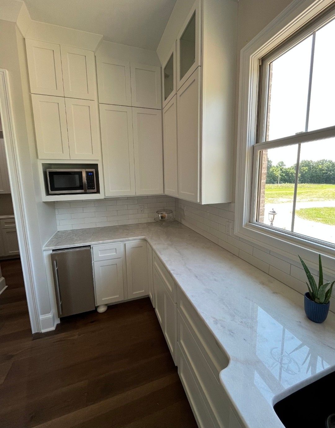 White kitchen cabinets with light countertop, window, built-in microwave, and stainless steel dishwasher.