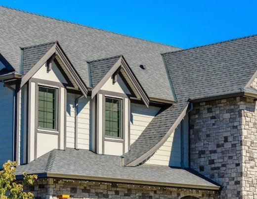 Stone and white house exterior with gray shingled roof and dormer windows under a blue sky