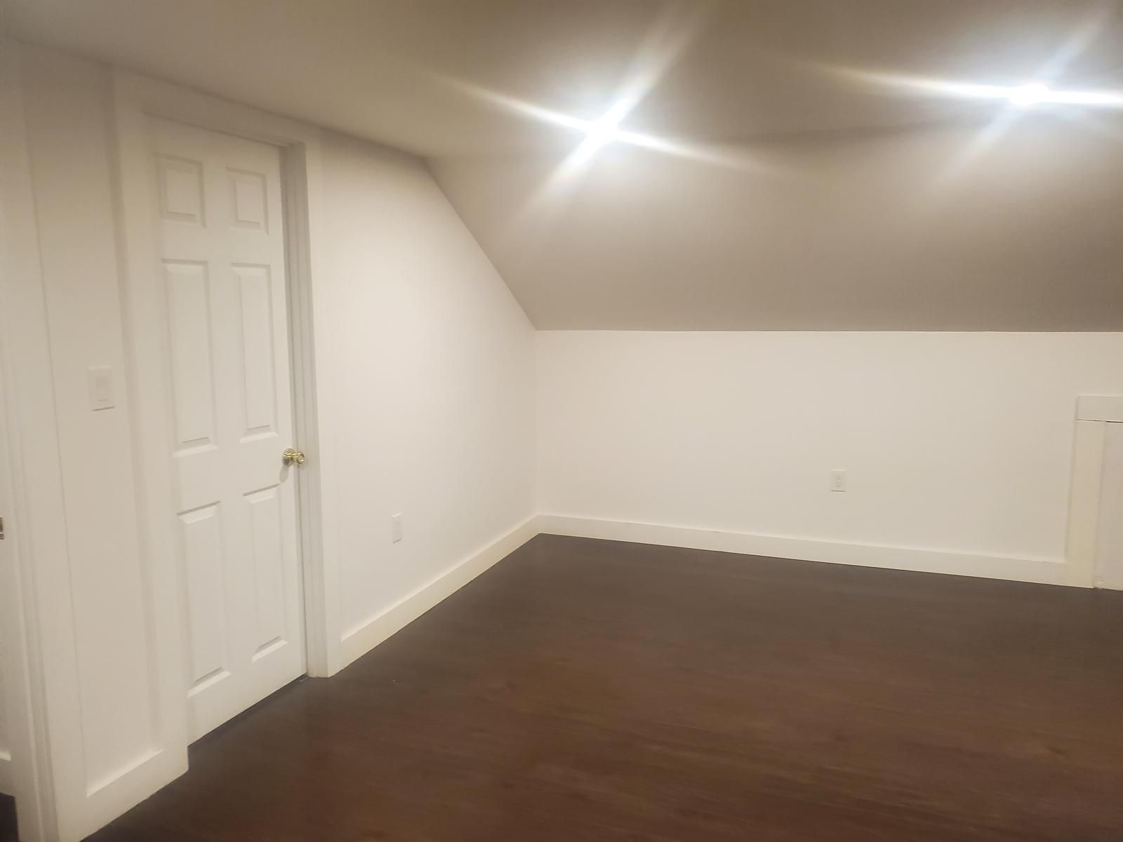Empty room with white walls, slanted ceiling, brown hardwood floor, and recessed lights