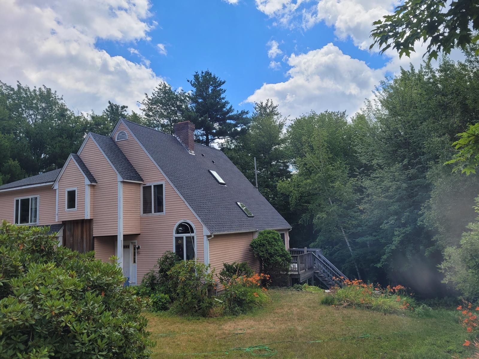 Brick house with steep roof and garden, surrounded by trees under a partly cloudy sky