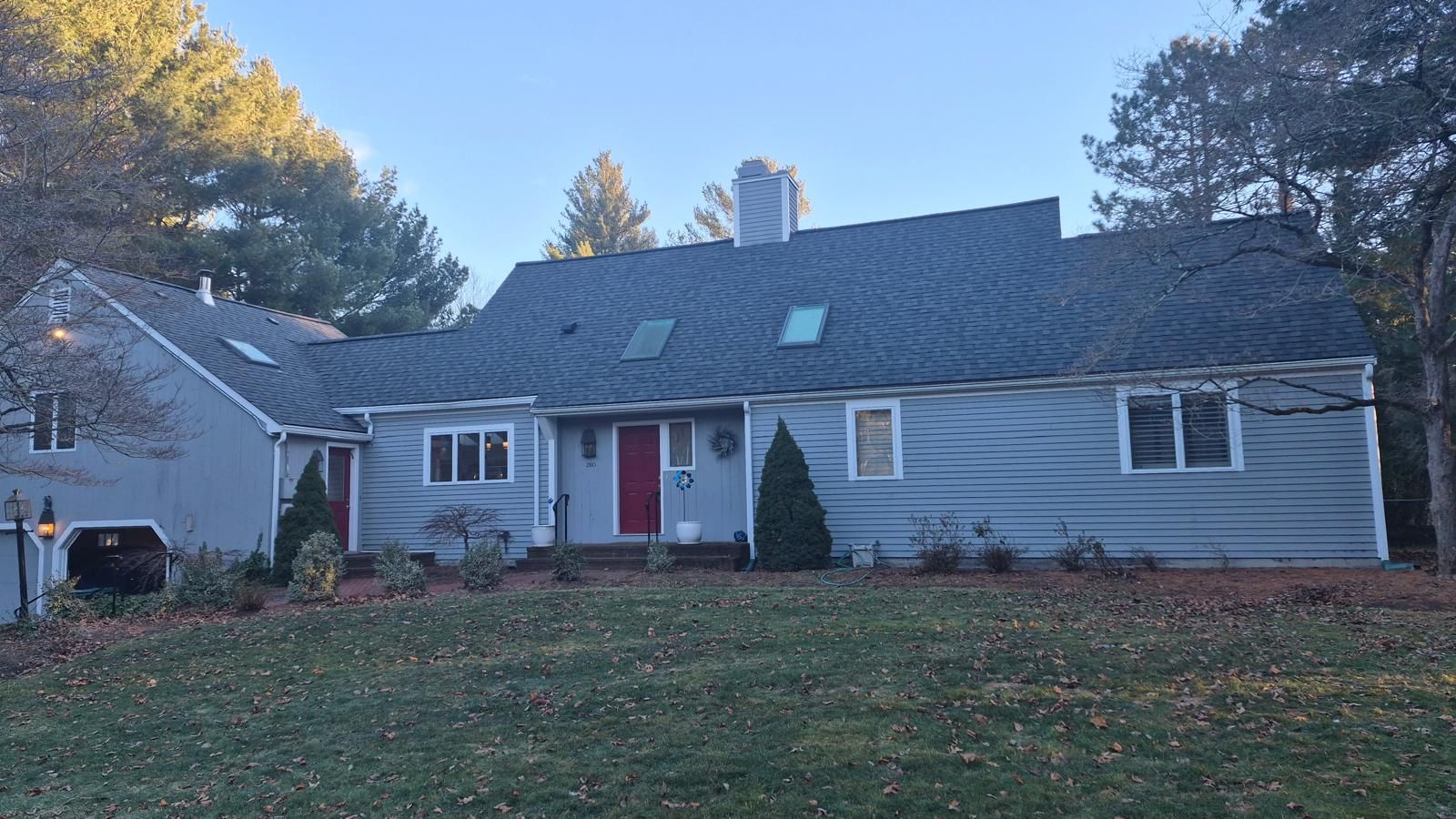 Gray suburban house with a red front door, dormer windows, and autumn trees in the yard